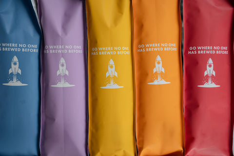 Five colorful pouches stacked horizontally, each in a different shade of red, orange, yellow, purple, and blue, featuring a white rocket illustration and the text 'GO WHERE NO ONE HAS BREWED BEFORE'.